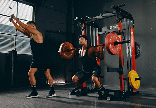 A man squats while another does overhead cable tricep extension on a power rack